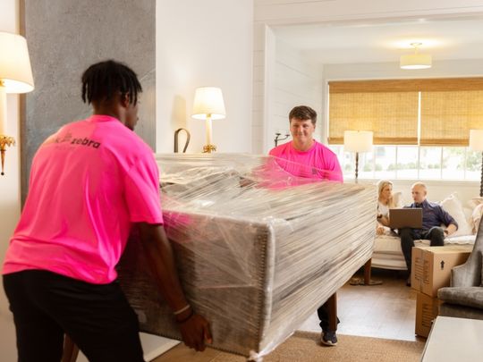 Two movers in bright pink shirts carry a plastic-wrapped sofa inside a modern home while a couple sits in the
background.